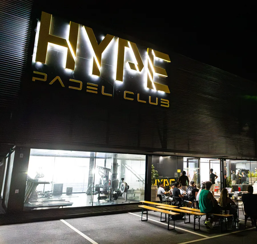 A fachada à noite, com o letreiro dourado Hype Padel Club aceso e gente na esplanada.